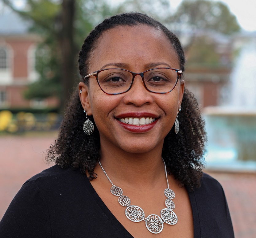 The face and shoulders of a woman with dark brown skin and glasses smiling directly at the camera. Blurred water fountain in the background.